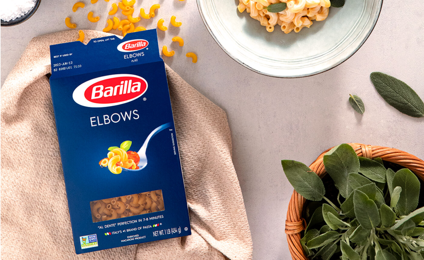 A box of Barilla elbow pasta rests on a beige cloth, accompanied by a bowl of steaming, cooked pasta and a basket filled with aromatic sage leaves atop a sleek gray surface.