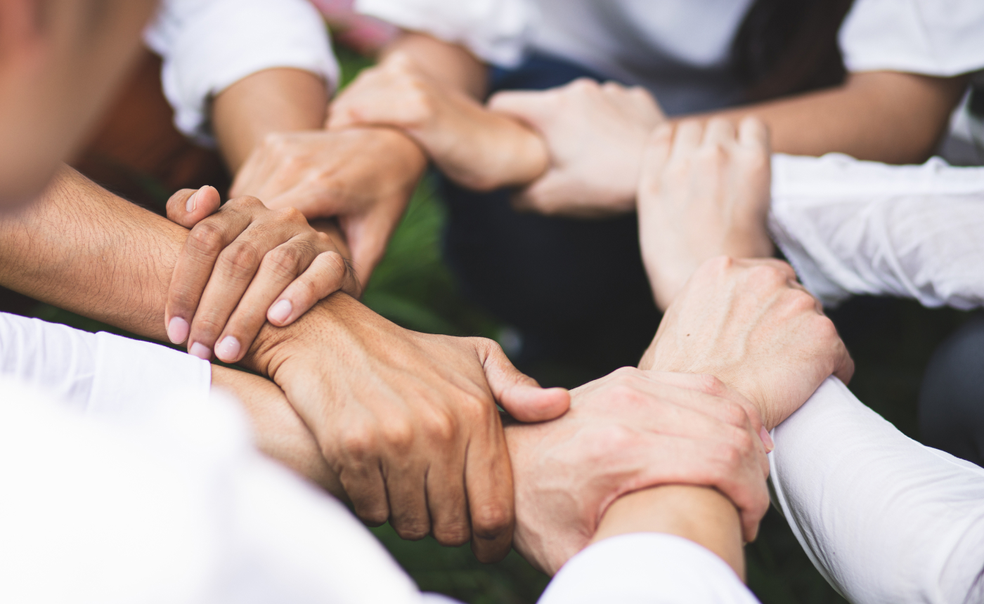A group of individuals forms a circle by holding each other's wrists, symbolizing unity and teamwork. Clad in white shirts, they stand on a field of vibrant green grass, emphasizing their connection to nature.