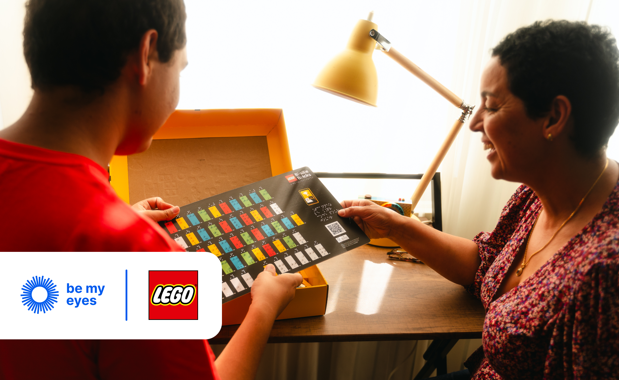 A person shares a LEGO instruction booklet with another at a desk lit by a lamp, while the LEGO and Be My Eyes logos are prominently displayed in the foreground.