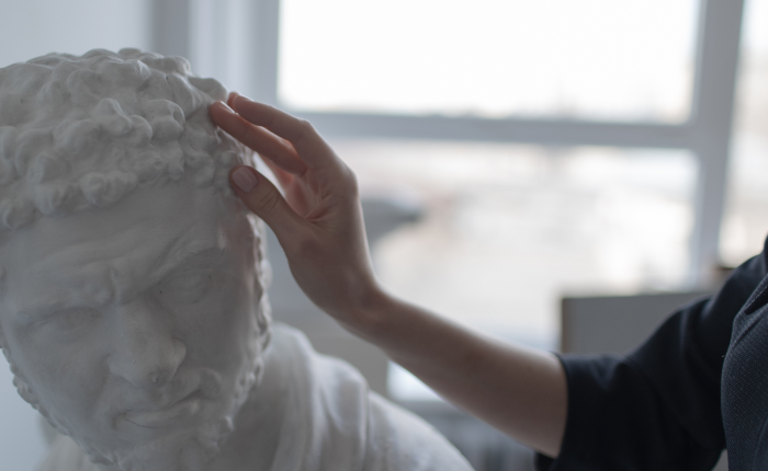 A hand gently touches the curly hair of a white marble bust in a softly lit room, where sunlight streams through the window.