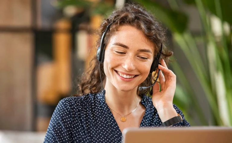 A woman with curly hair, wearing a headset, cheerfully gazes at her laptop in an indoor setting adorned with lush plants.