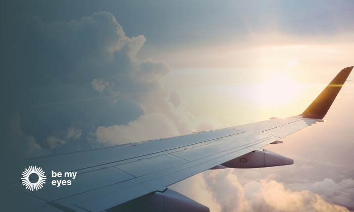 Travel and hospitality image showing an airplane wing against a backdrop of clouds and sunlight, viewed from the passenger window, with the "Be My Eyes" logo in the corner.