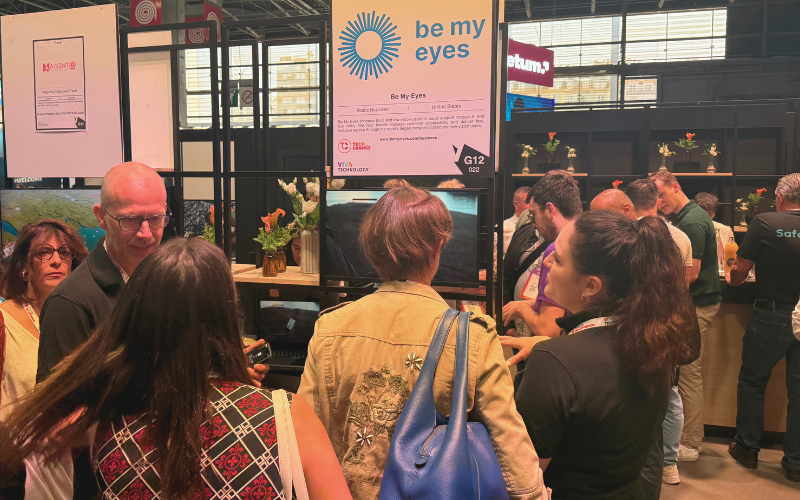 A group of people stand in front of a "Be My Eyes" booth at VivaTech 2025 with informational signs and displays visible in the background.