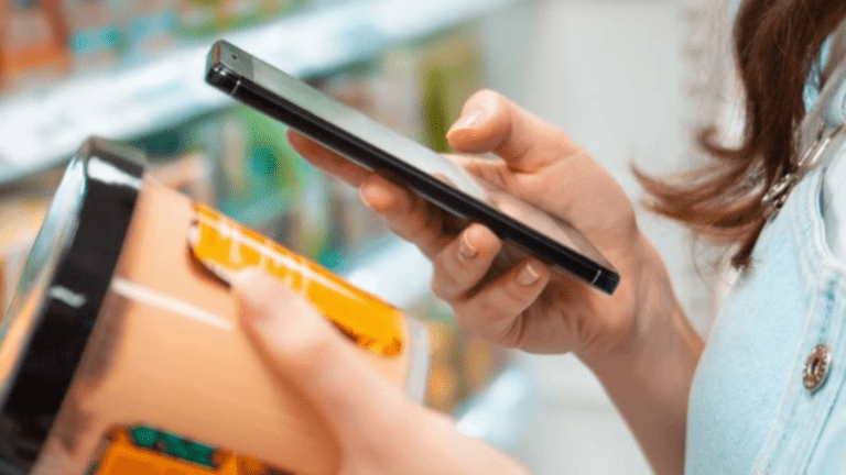 A person holds a packaged product in one hand and a smartphone in the other, possibly scanning the barcode or searching for information in a store aisle while wearing smart glasses.