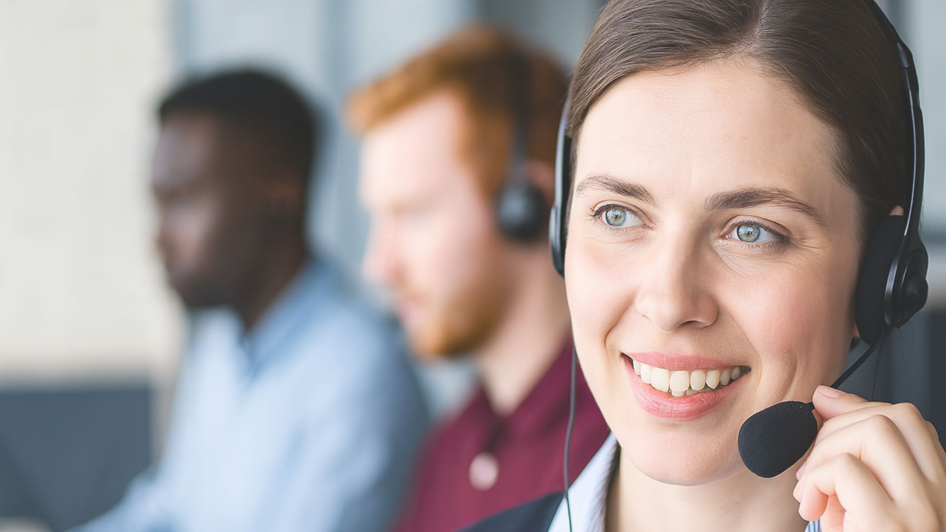 A woman wearing a headset smiles at the camera, showcasing accessible support, while two coworkers in headsets work at computers in the background.