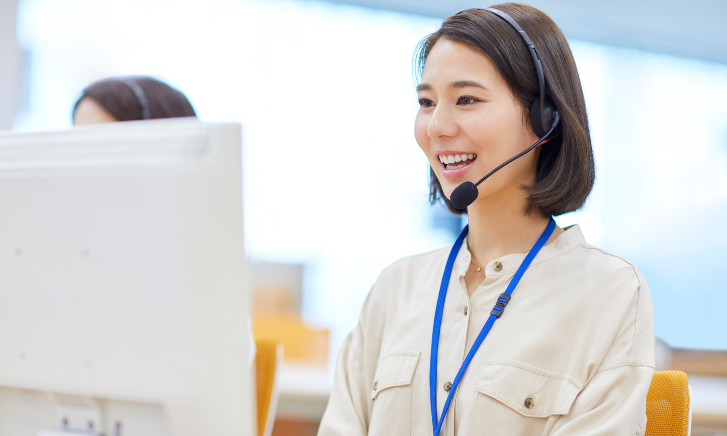 A woman wearing a headset and lanyard sits at a desk, smiling while working on a computer in an office environment