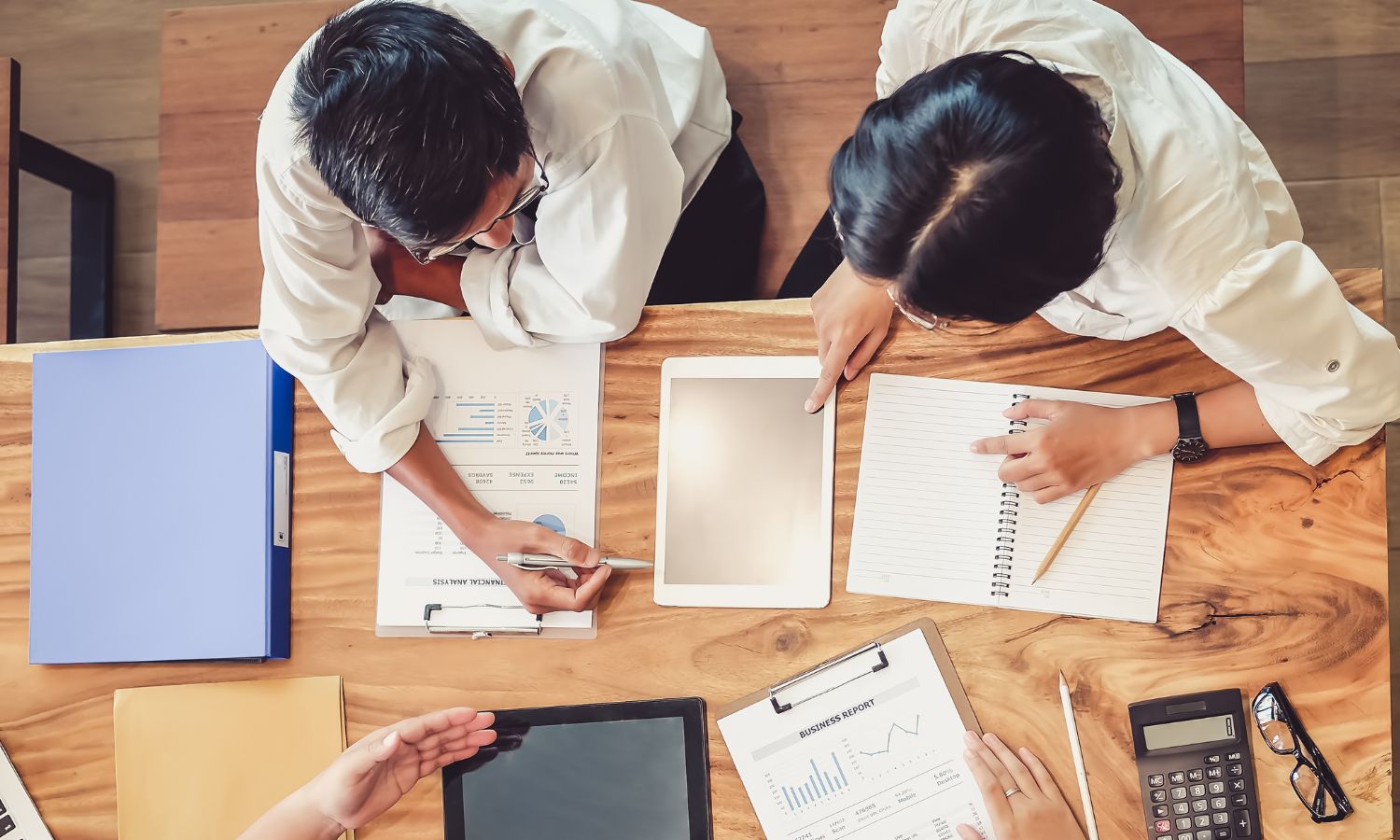 Two people sit at a wooden table reviewing charts, graphs, and notes, discussing KPIs, with documents, a calculator, and tablets spread out in front of them.