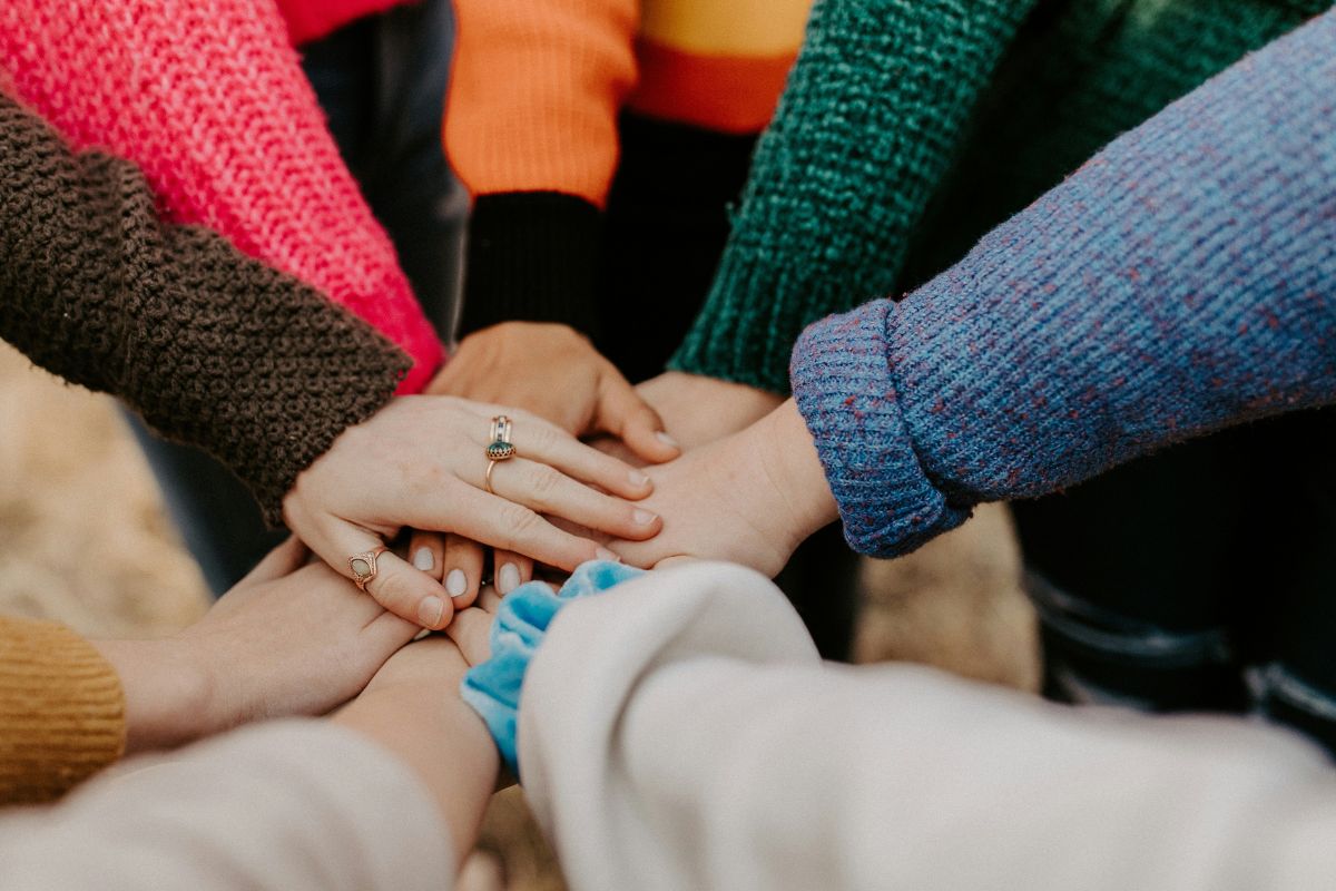 A group of people wearing colorful sweaters stand in a circle with their hands stacked together in the center, embodying how corporate volunteering strengthens brand trust through unity and teamwork.