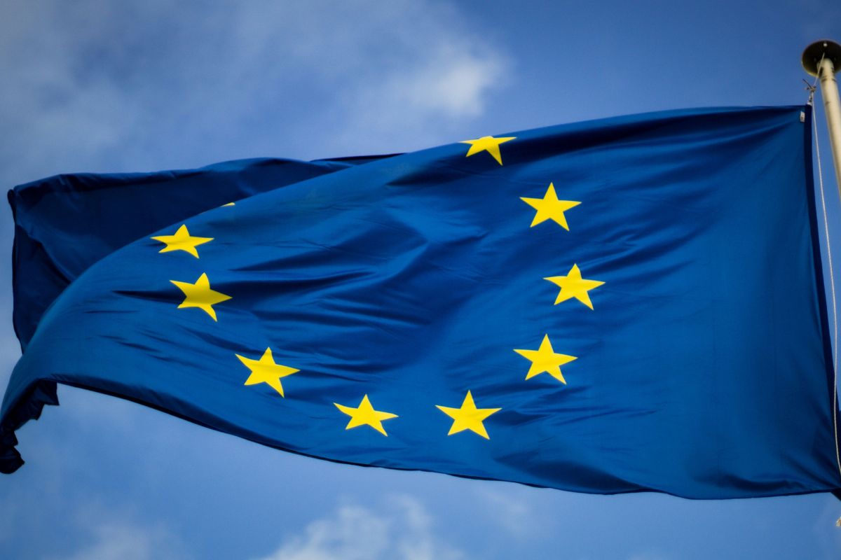 The European Union flag with twelve yellow stars in a circle on a blue background is shown fluttering against a partly cloudy sky.