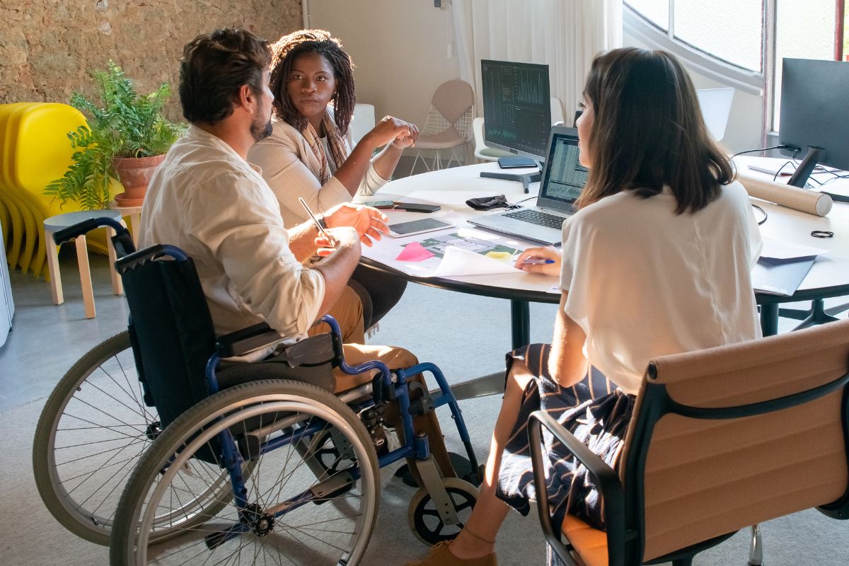 Three people, including a man in a wheelchair, sit around a table with laptops and documents, having a discussion in a modern office
