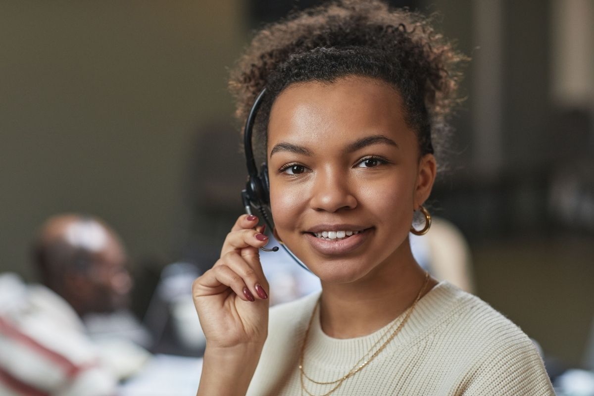 Young woman wearing a headset and smiling, sitting indoors, possibly working in a customer service or call center environment