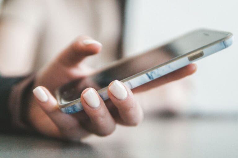 A person with manicured nails holds a smartphone in one hand over a smooth surface.