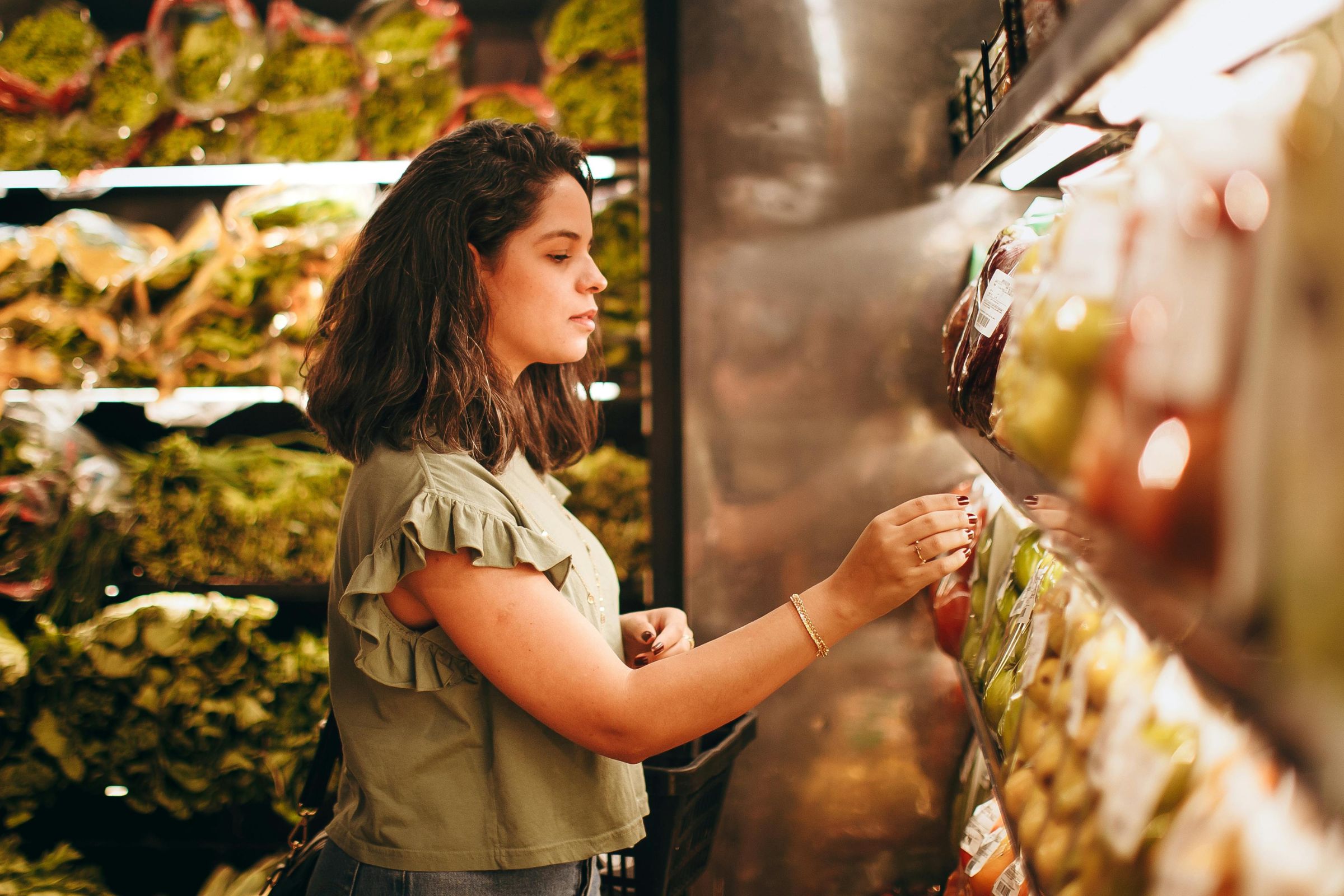 A woman stands in a grocery store produce section, selecting items from a shelf lined with packaged fruits and vegetables