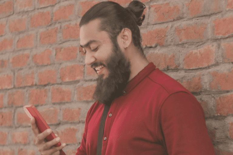 A man with a beard and a bun, wearing a red shirt, stands against a brick wall, smiling at his phone.