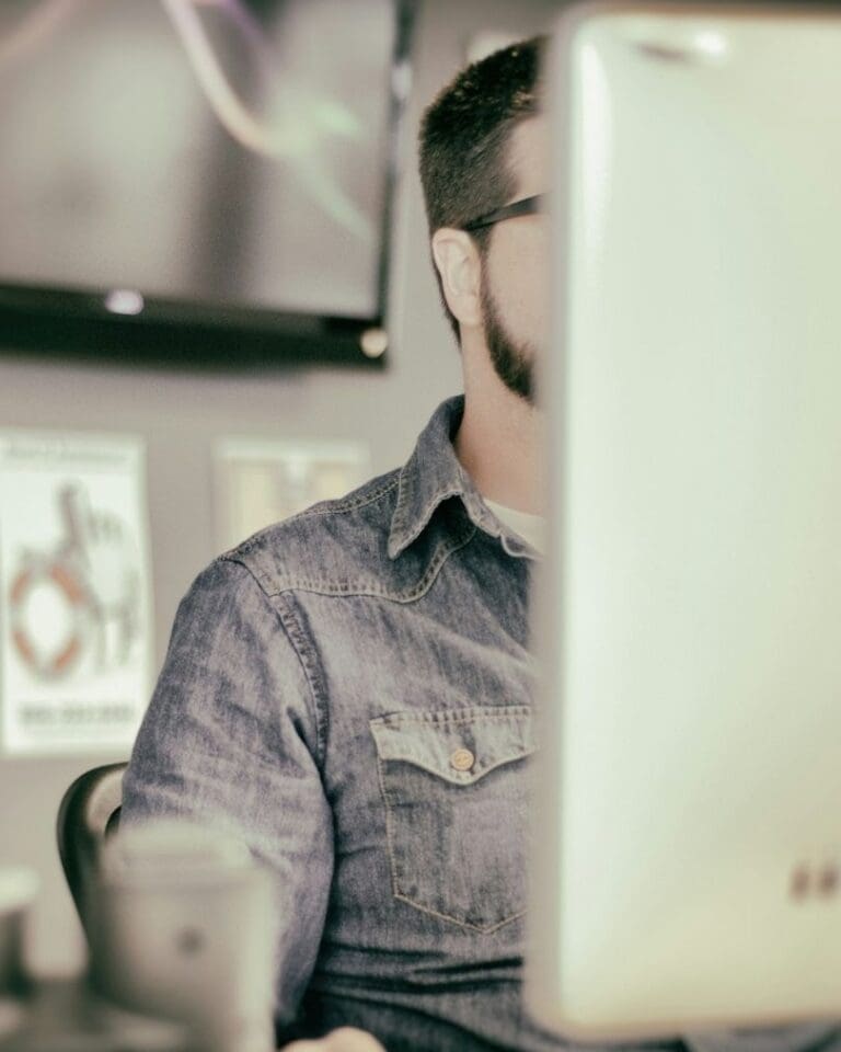 A man with a beard and glasses works at a computer in an office, partially obscured by a monitor. Posters are visible on the wall in the background.