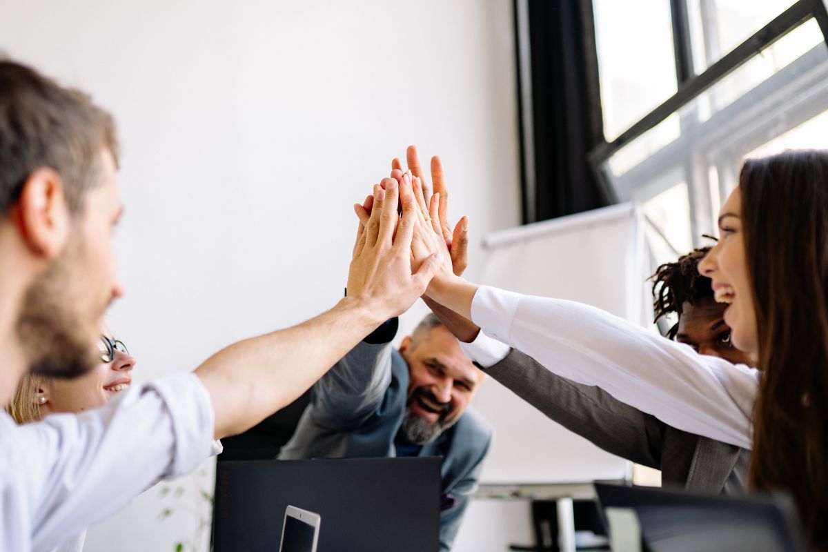 A group of coworkers in an office raising their hands together in a group high-five. Several hands meet in the center of the photo, forming a stacked high-five. On the right, a woman with long dark hair smiles, wearing a white top. On the left, a bearded man in a light shirt is in the foreground, slightly out of focus. In the background, an older man with gray hair and a gray beard smiles, leaning forward behind a laptop. Another person with dark skin and short dreadlocks is partly visible behind the woman on the right. A bright window and a whiteboard or flip chart stand are in the background, suggesting a meeting or teamwork moment. Described with Be My AI
