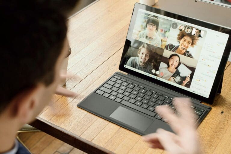 A tablet-style laptop on a wooden table showing a video call with four people in a grid. The screen also has a chat panel on the right with small messages and emoji reactions. In the foreground, a person wearing glasses is turned toward the screen, with a raised hand blurred in motion. The device has a black keyboard attached, and the camera angle is slightly above and behind the foreground person. Described with Be My AI