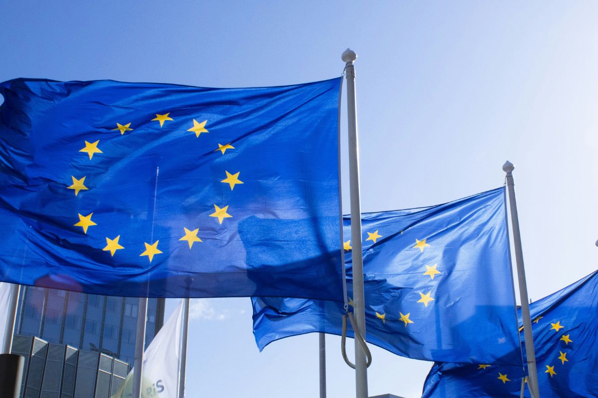 Three European Union flags with yellow stars on a blue background are waving in the wind against a clear blue sky, with buildings partially visible in the background.