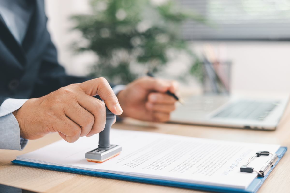 A person stamps a document on a clipboard at a desk, with a laptop and pen in hand.