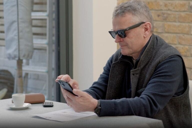 A man wearing sunglasses sits at a table with a smartphone, cup of coffee, and papers in front of him, next to a window and a brick wall.