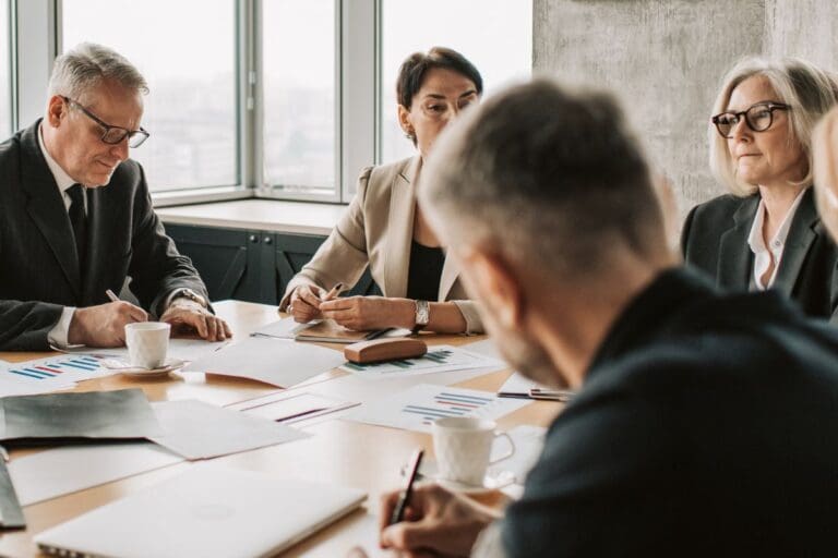 Four people in business attire sit around a conference table with documents, charts, and coffee cups.