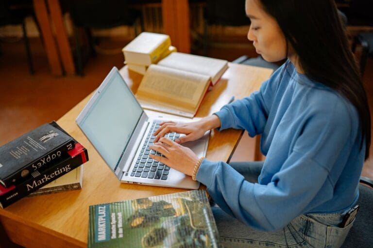 A person in a blue sweater types on a laptop at a wooden table, surrounded by open books and textbooks.