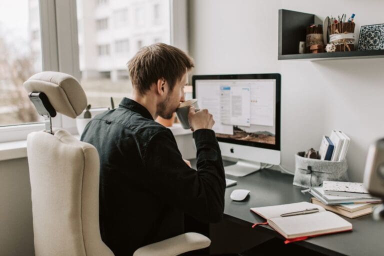 A person sits at a desk, drinking from a mug while working on a computer with multiple windows open. Office supplies and a notebook are on the desk.