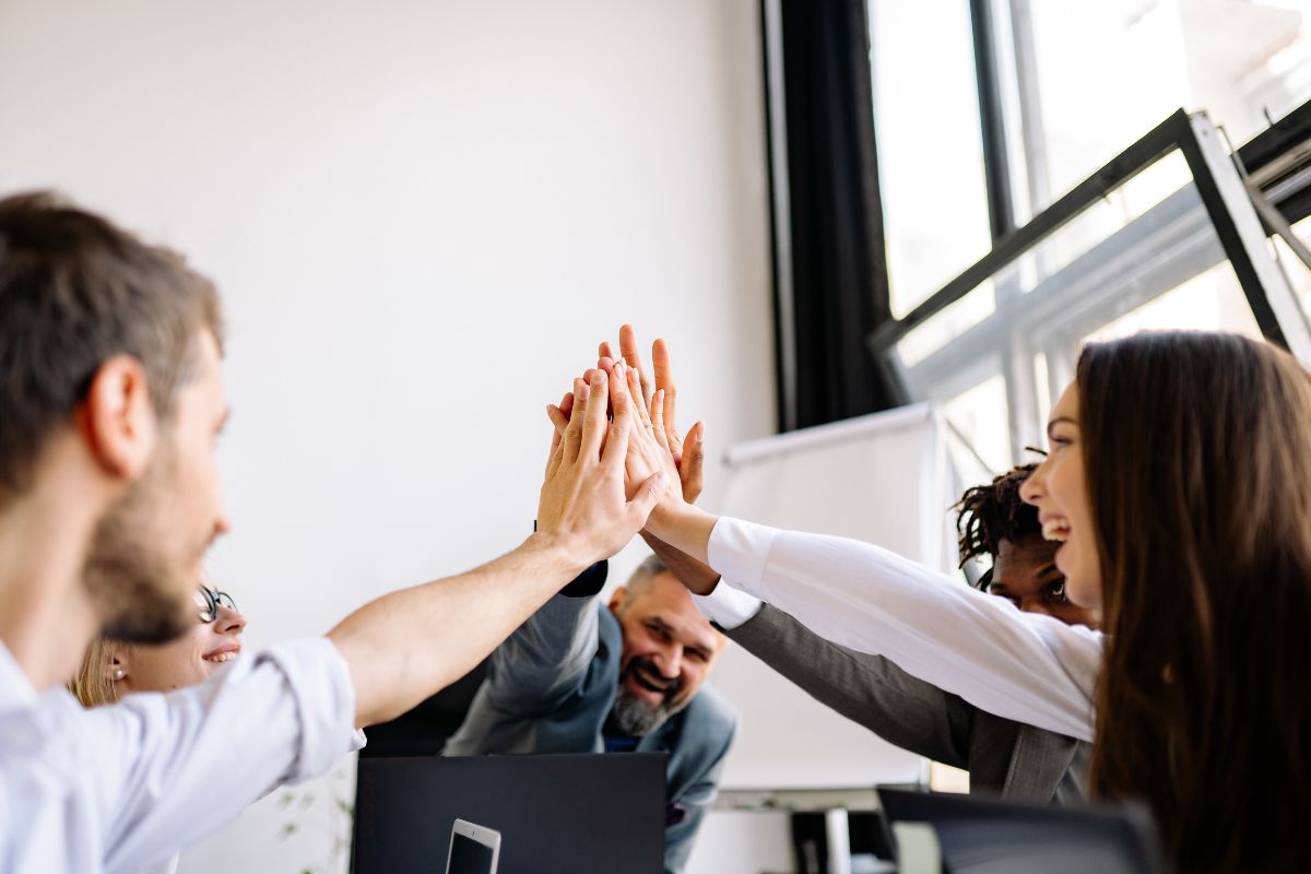 A group of people in business attire smiles and gives a group high-five in a bright office setting.