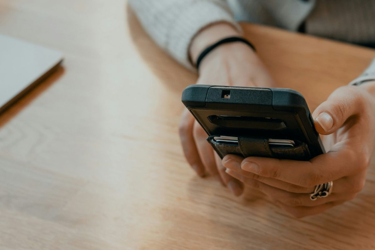A person holding a smartphone with both hands, seated at a wooden table, follows accessibility standards, with part of a laptop visible in the background.