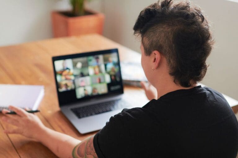 Person with short hair sits at a wooden table using a laptop for a video conference, utilizing service AI tools, with a notebook and pen nearby.