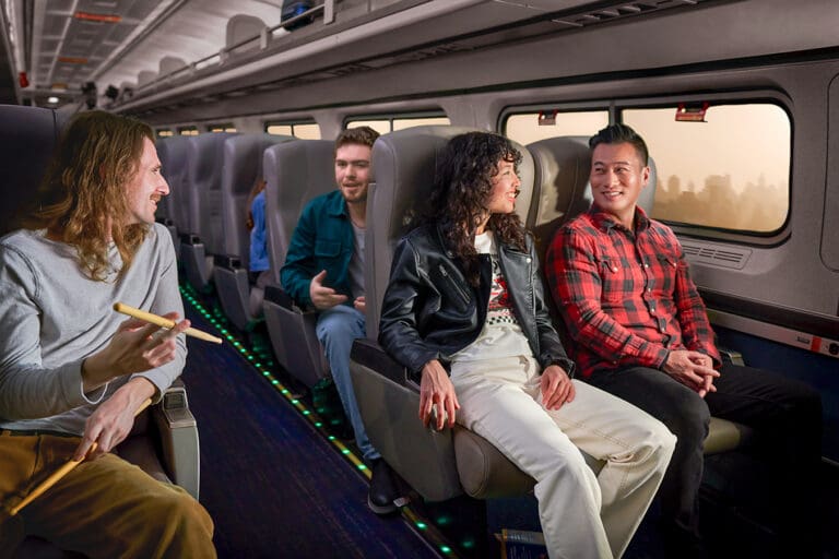 Four people sit and talk on a train with city buildings visible through the windows. The seats are arranged in pairs, and overhead lights illuminate the cabin.