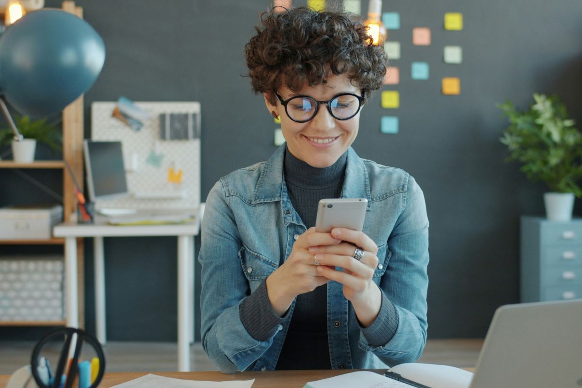 A person with short, curly brown hair and light skin is sitting at a desk, smiling while using a smartphone. The person is wearing round black glasses, a gray turtleneck, and a light blue denim jacket. Both hands are holding the phone, and the person’s attention is focused on it, with a relaxed, content expression. On the desk in front of them are papers, a pen holder with pens, and the corner of an open laptop. In the background there is a dark wall with many colorful sticky notes, a lit hanging bulb, a potted plant on the right, and a white desk with a monitor and other office items on the left, giving the space a modern, casual office or home-office feel. Described with Be My AI.