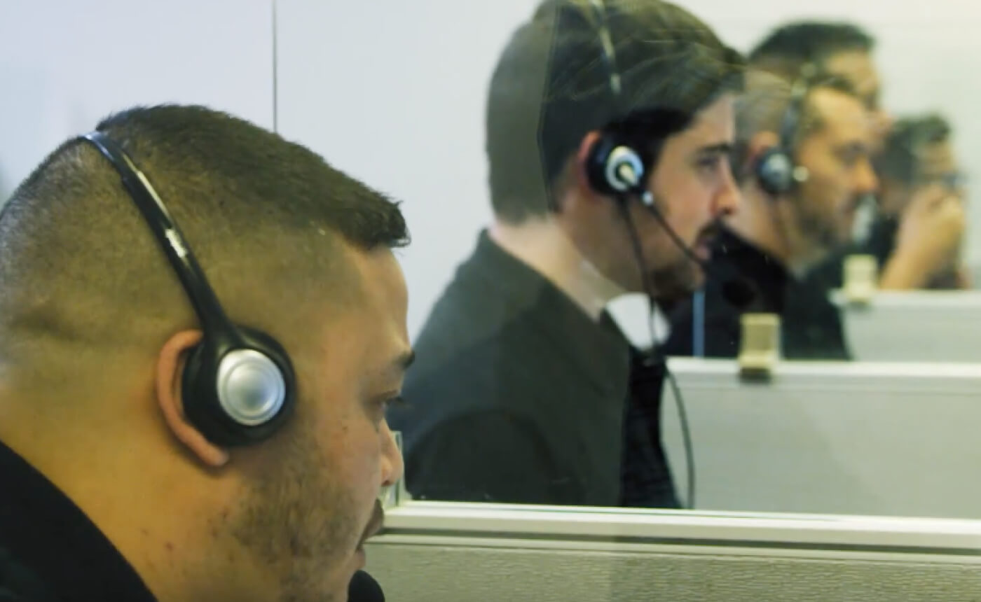 Three men wearing headsets are diligently working at individual cubicles, separated by glass dividers. The focus in this bustling environment is evident as they manage tasks efficiently, embodying teamwork and communication skills.