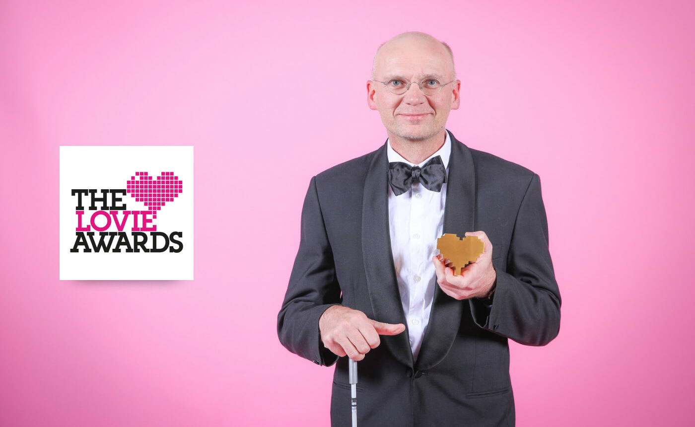 A person in a tuxedo holds a heart-shaped award in front of a pink background, standing next to a sign that reads "The Lovie Awards.