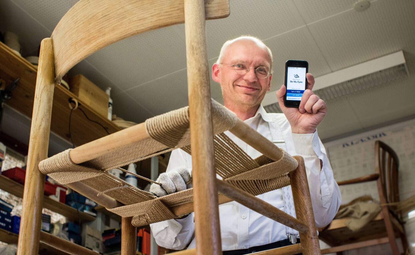 In a busy workshop, someone adeptly restrings a wooden chair while holding up a smartphone. Various tools and materials are scattered in the background, highlighting the craft in progress.