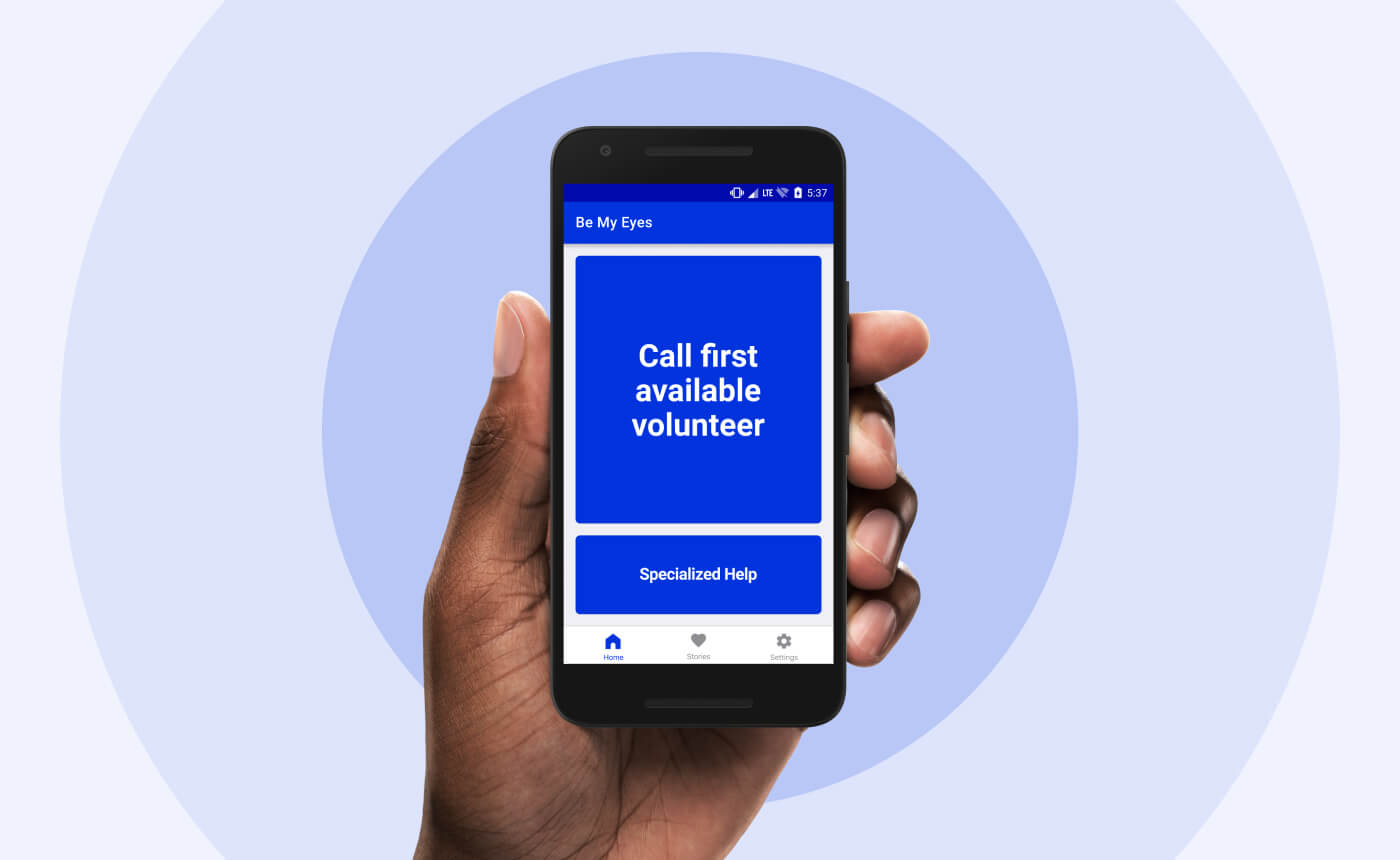 A hand holds a smartphone displaying the "Be My Eyes" app. The screen features a blue button labeled "Call first available volunteer" and an option for "Specialized Help" at the bottom, making it simple to connect for assistance.