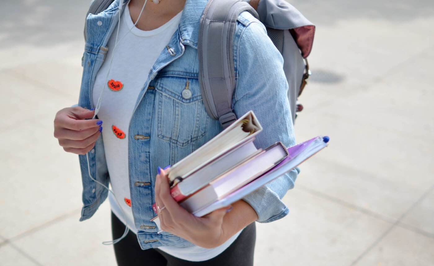 A person sporting a denim jacket and headphones clutches books and a backpack, confidently standing on the bustling sidewalk.