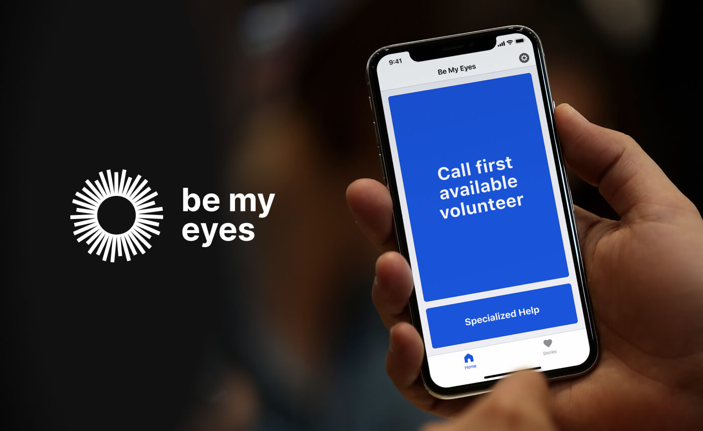 A person holds a smartphone featuring the "Be My Eyes" app, showcasing "Call first available volunteer" on the screen. The app's logo is prominently displayed on the left side, highlighting its accessibility feature.