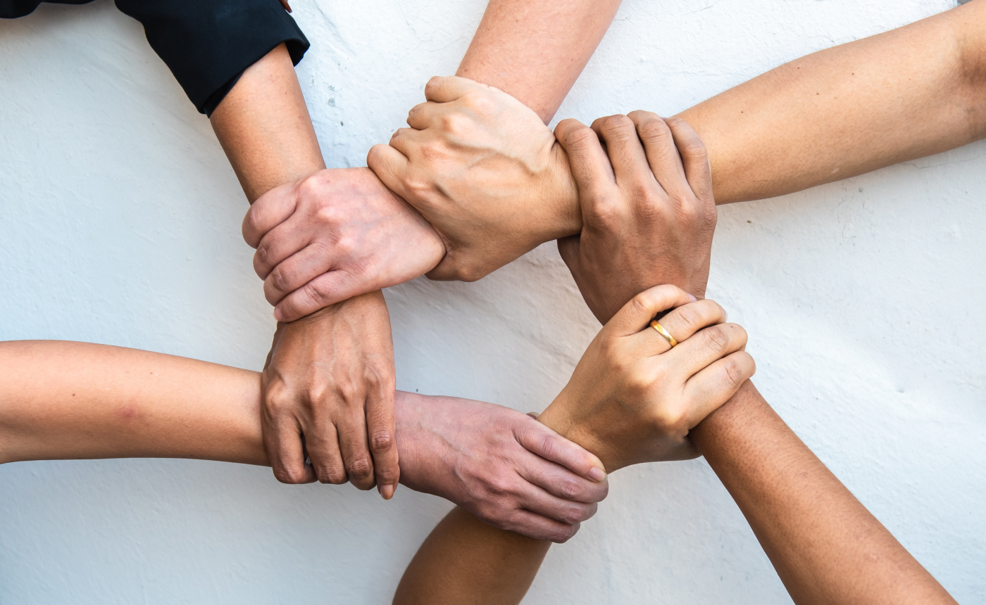 Six hands of diverse skin tones are linked in a circular formation, each gripping the wrist of the next. This powerful image symbolizes collaboration while reflecting an unbreakable bond and collective strength and resilience.