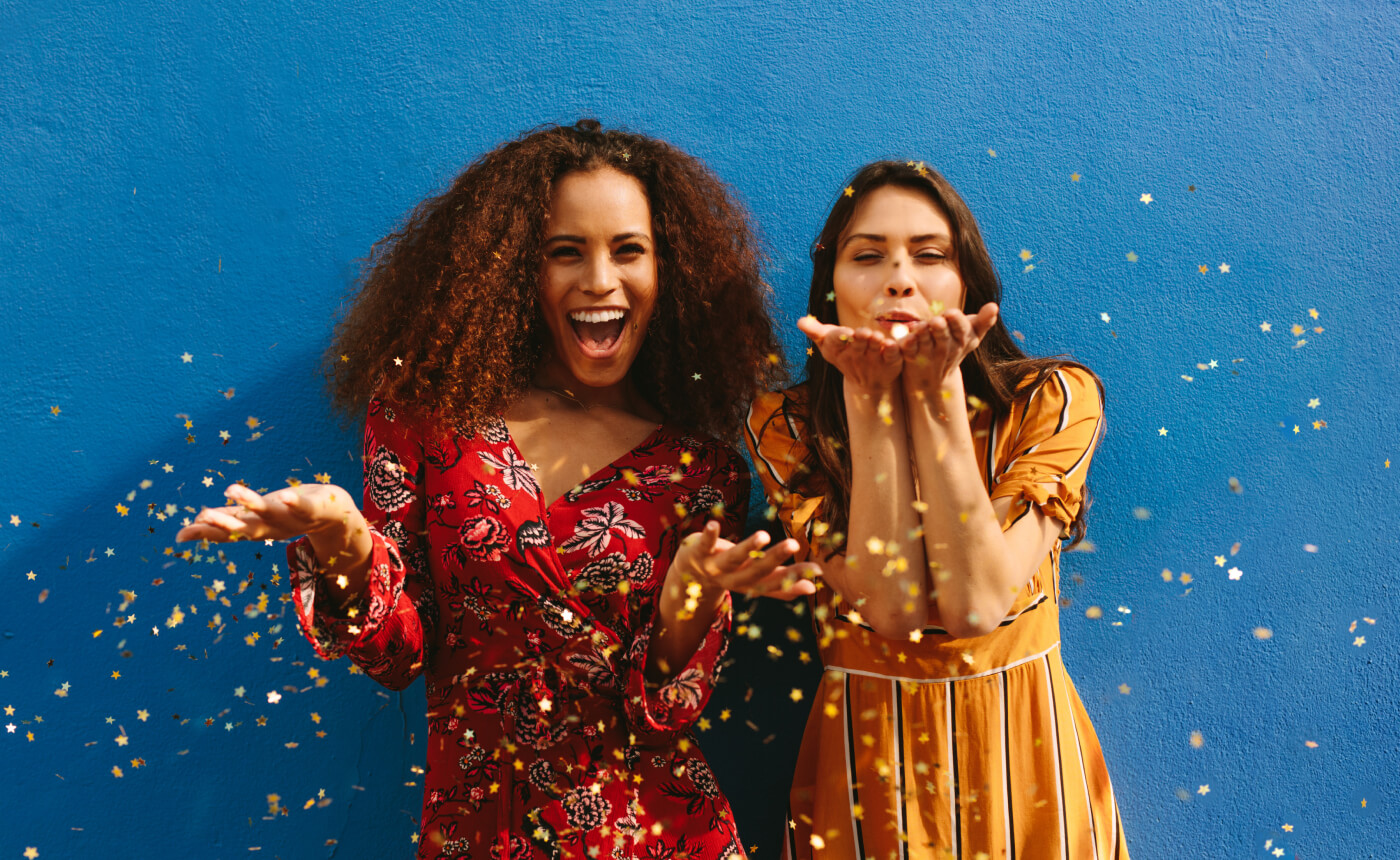 Two people stand against a blue wall, smiling and blowing gold confetti. One wears a vibrant red floral dress, while the other shines in a lively yellow striped dress, capturing a moment of pure joy.