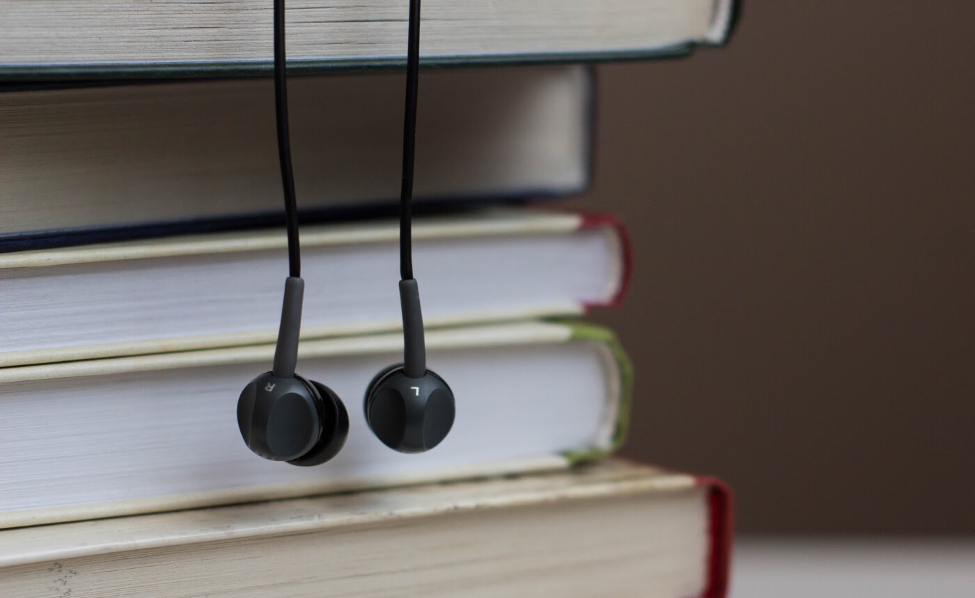 Black earphones lie against a stack of hardcover books, their pages and spines clearly visible on the flat surface. Whether you're diving into an audiobook or enjoying your favorite tunes, these earphones offer a perfect blend of tech and literature.