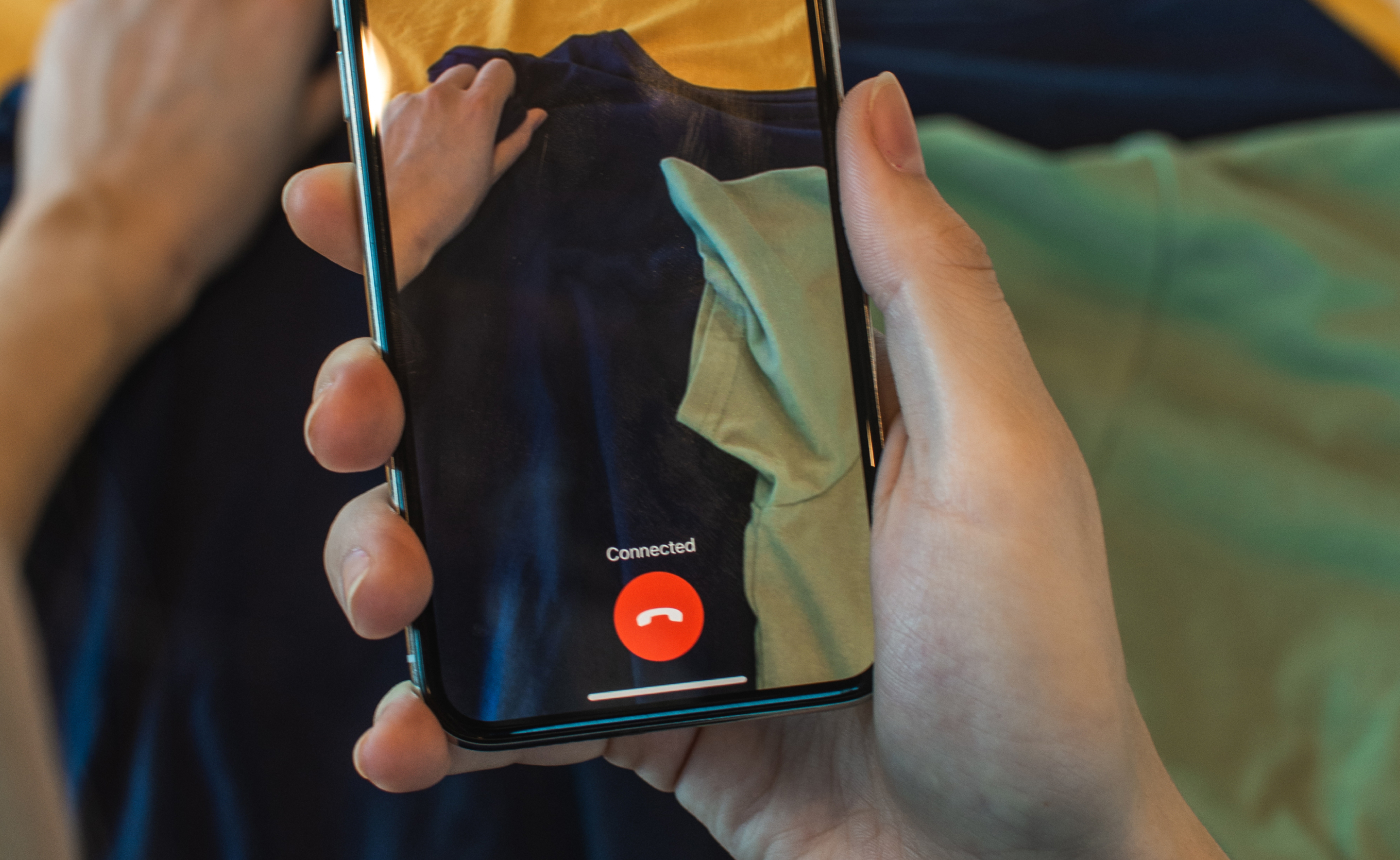A person holds a smartphone showing a video call screen. A red button labeled "Connected" stands out, while their other hand, clad in a blue garment, is visible.
