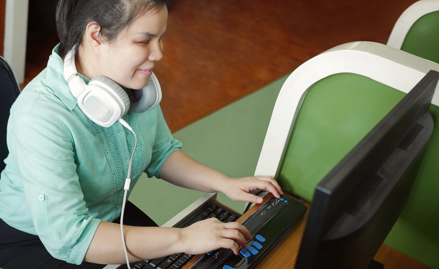 A person seated at a desk in an office environment uses a computer with a Braille display while wearing headphones, efficiently navigating through tasks.