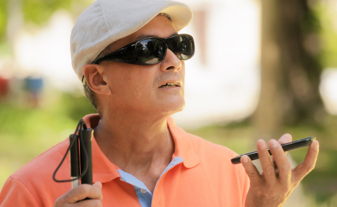 A man wearing sunglasses and a cap skillfully navigates an outdoor setting, using a smartphone with one hand while holding a white cane in the other.
