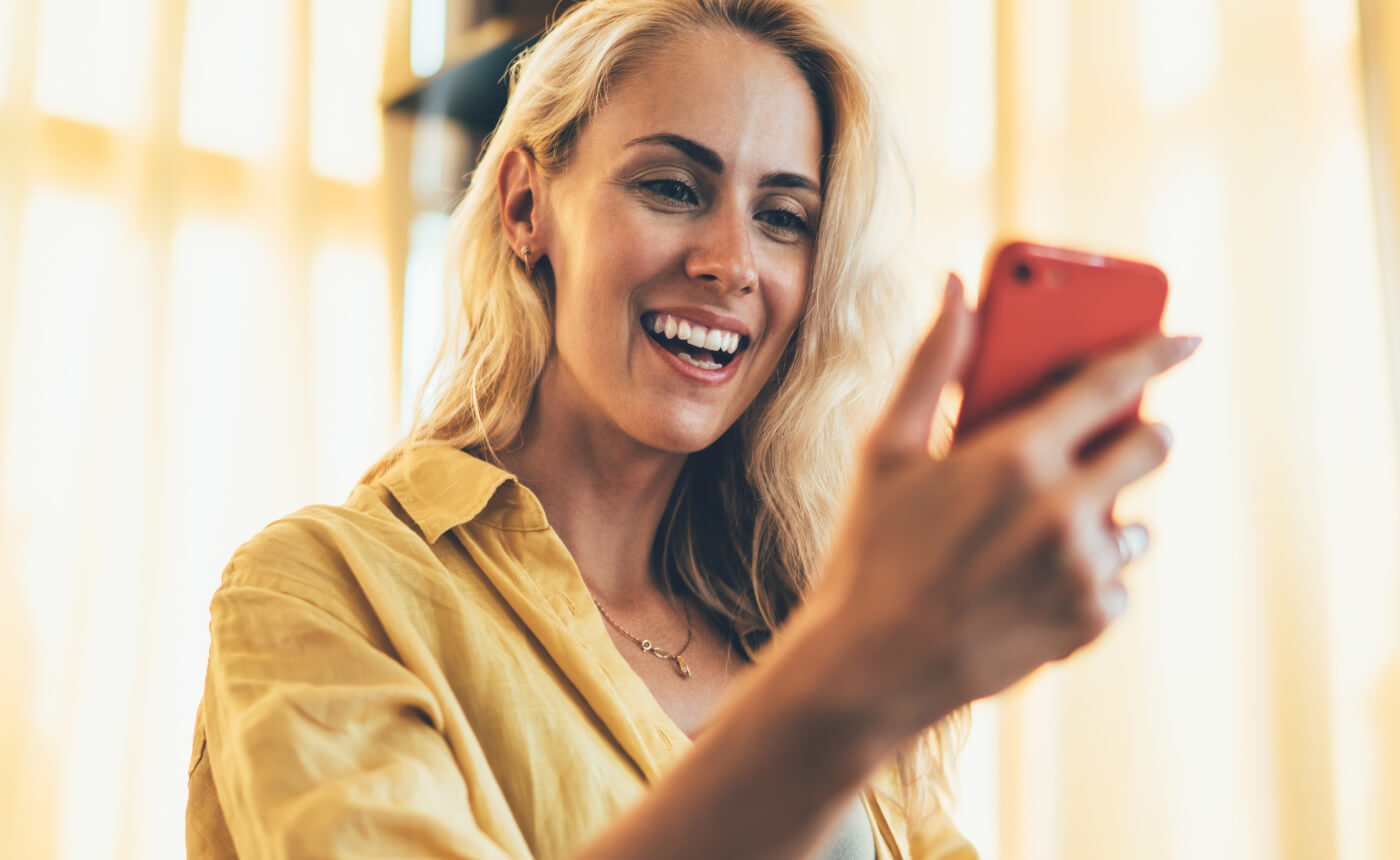 A woman in a yellow shirt smiles brightly as she reads an uplifting message on her phone.