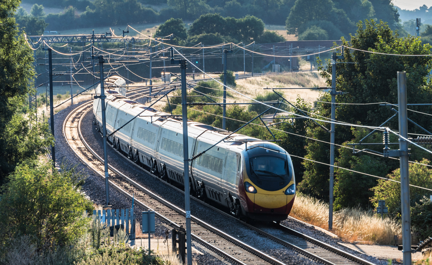 A high-speed passenger train glides swiftly along a curved railway track, cutting through the serene rural landscape dotted with trees and crossing beneath overhead power lines.
