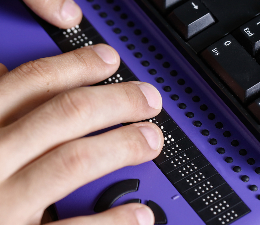 Close-up of a person's hand reading braille on an electronic braille display with a purple surface, adjacent to a keyboard.