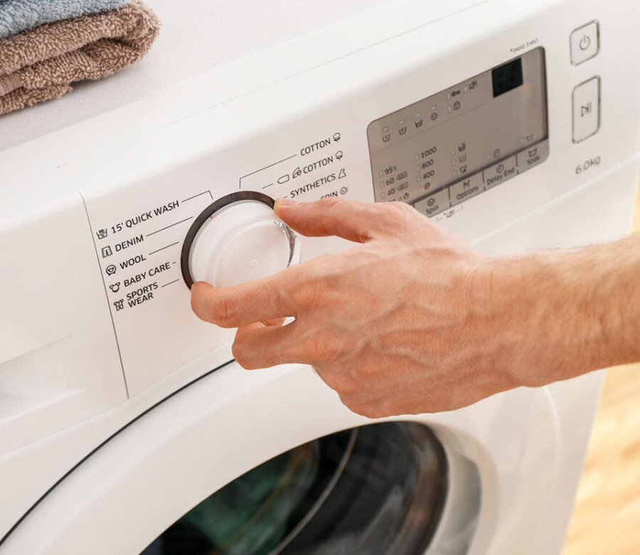 A person adjusts the dial on a washing machine, with various wash settings visible, including cotton, wool, and baby care.