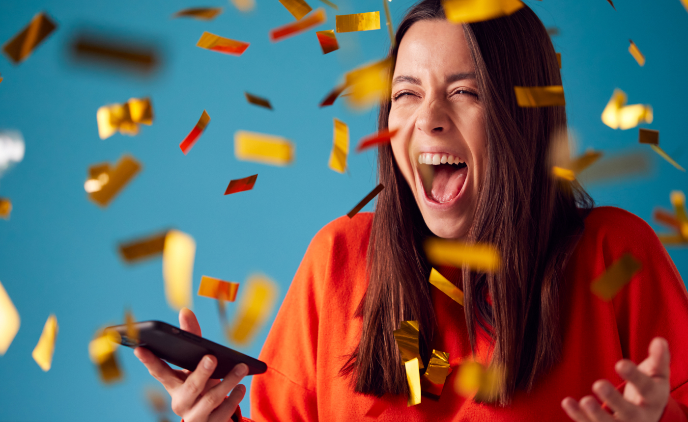 A woman in a red shirt is excitedly holding a smartphone as gold and red confetti rains down around her against a vibrant blue background, capturing a moment of pure joy.