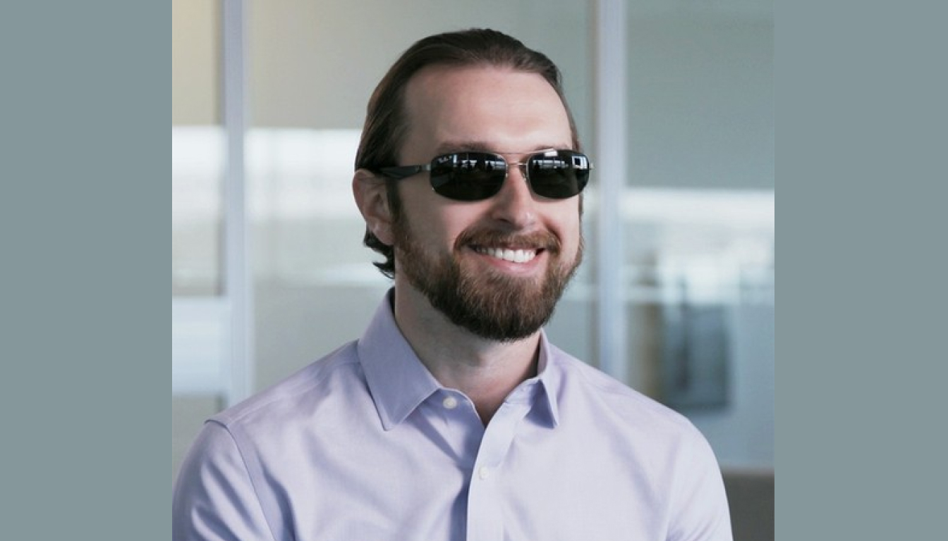 A man with a beard and a ponytail is wearing dark sunglasses and a light-colored shirt, smiling in a bright room with glass partitions.